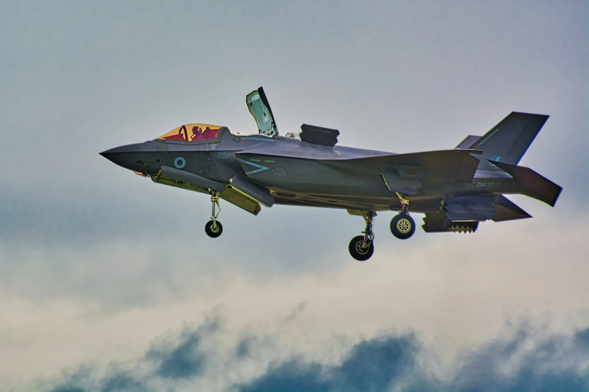 A grey fighter jet in flight with its landing gear down against a cloudy sky