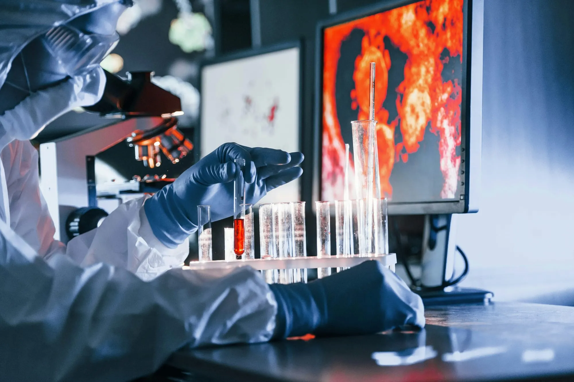 A gloved scientist in protective gear holding a rack of test tubes in front of a monitor